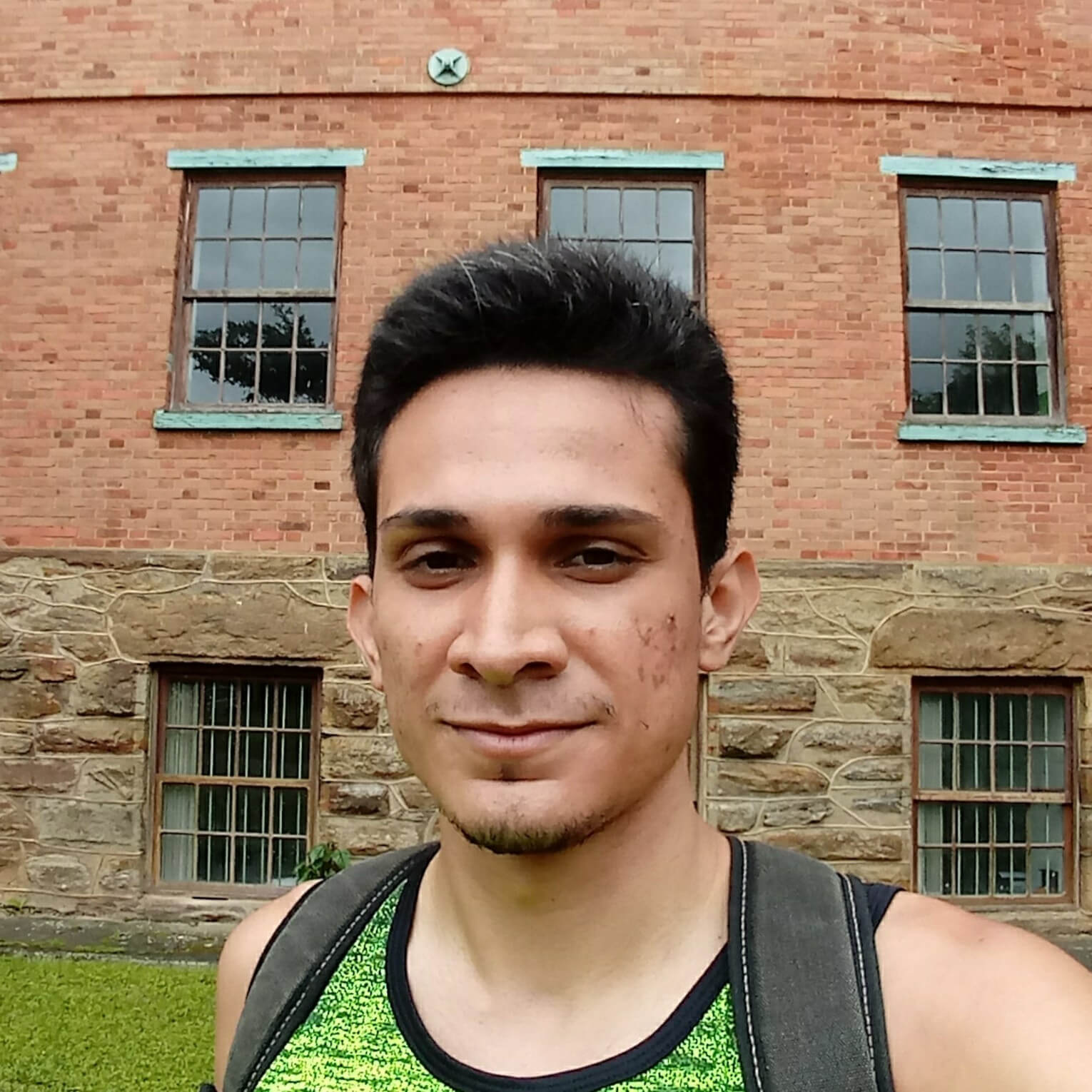 Gabriel, a white man with short dark hair, wearing a green t-shirt and a backpack, standing in from of a campus